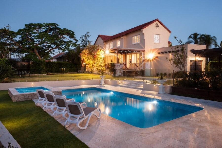 A well-illuminated backyard featuring a swimming pool in the foreground and a home in the background.