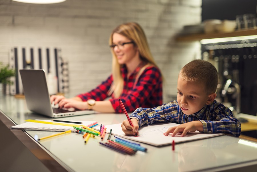 A woman working on the computer on the kitchen counter while her son colors next to her.