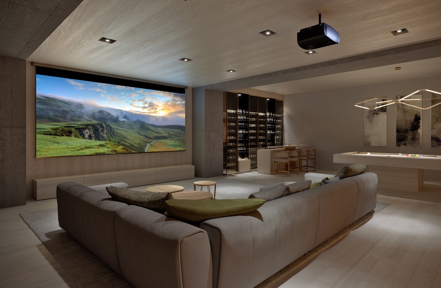 A modern home theater/media room with a large screen, projector, sofa seating, and a pool table in the background.