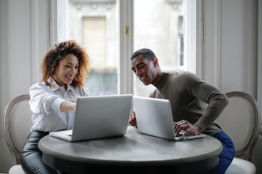 A man and a woman work on their laptops. The woman is pointing at her screen while the man looks over.